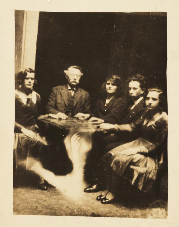 a sepia image of five adults at seance table with ectoplasm streaming below a table